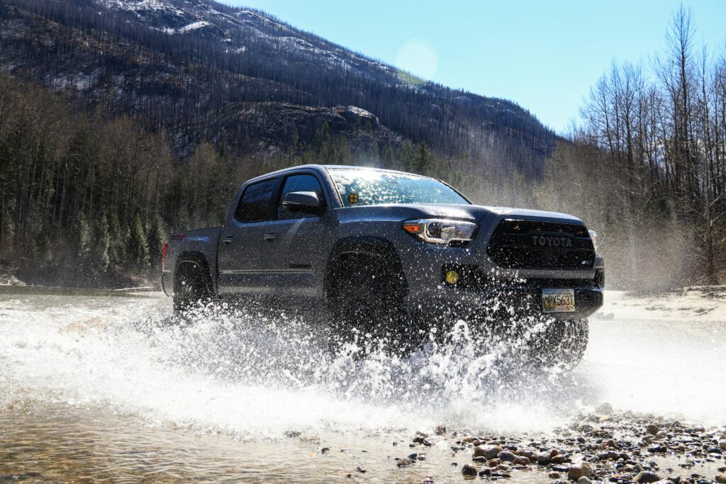 A Toyota Tacoma drives through a water puddle with splashing effects in a rugged outdoor setting.